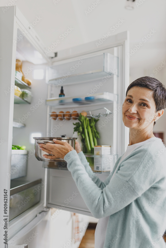 Side view of charming mature woman housewife putting soup in refrigerator to storage till next day to save food, holding metallic saucepan in hands and smiling at camera. Domestic life concept Side view of charming mature woman housewife putting soup in refrigerator to storage till next day to save food, holding metallic saucepan in hands and smiling at camera. Domestic life concept