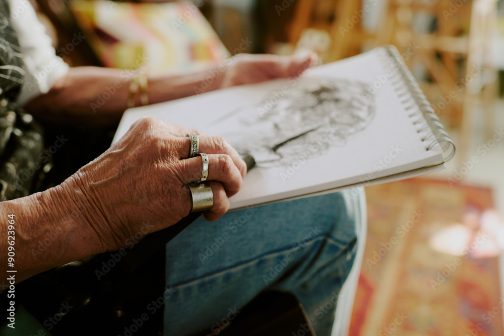 Obraz premium Closeup of wrinkled hands of unrecognizable senior female artist drawing portrait in sketchbook with charcoal pencil