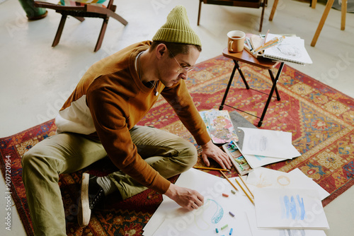 High angle view of young Caucasian man sitting on vintage carpet drawing picture on paper with crayons photo