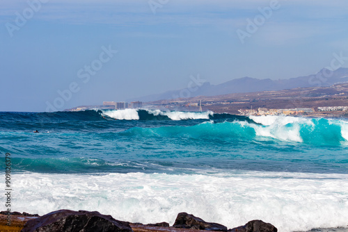 Tenerife, Spain 02 11 2013:  Young man surfing on a big wave with white foam doing radical maneuvers on the wave.  Mountains and village in the background with hotels. Blue sky. Surfing day. A