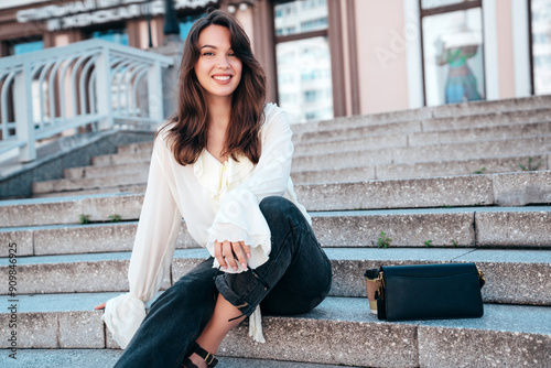 Young beautiful smiling hipster woman in trendy summer white blouse and jeans. Carefree woman posing in the street in sunny day. Positive model outdoors. Cheerful and happy. Sits at stairs