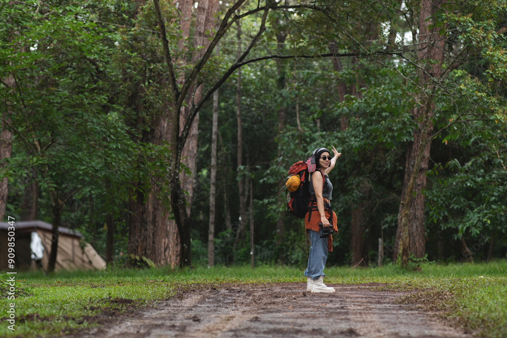 Fototapeta premium Asian female backpacker walks through pine forest to her campsite, raised hand to show her success amidst the small raindrops, journey with backpack concept