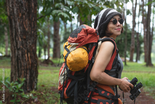 Portrait of Asian female backpacker holds binoculars to view natural scenery and birds while hiking through pine forest, journey with backpack concept