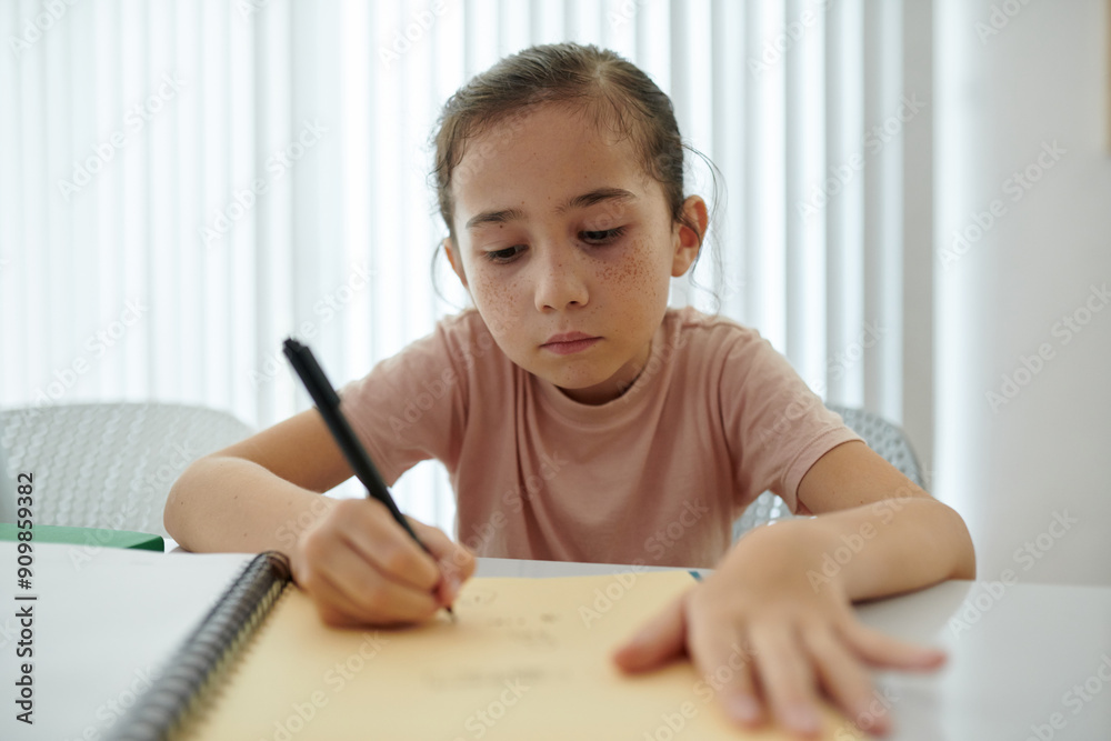 Young girl with focused expression drawing in notebook at table in bright room with white background emphasizing her concentration and creativity Young girl with focused expression drawing in notebook at table in bright room with white background emphasizing her concentration and creativity