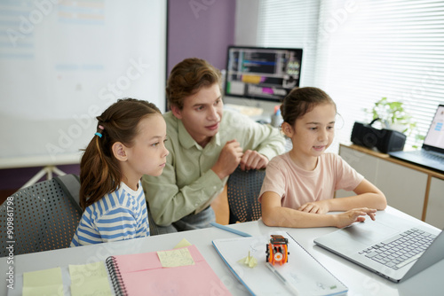 Teacher helping two young children with computer programming project while they are seated at desk. Classroom has educational materials and computer equipment in the background photo