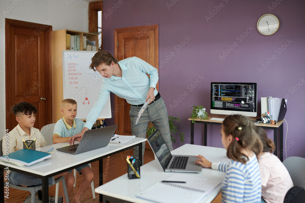 Teacher assisting young students with their laptop tasks in a classroom setting with educational materials Teacher assisting young students with their laptop tasks in a classroom setting with educational materials