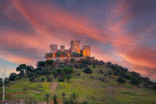 The ancient Almodovar del Rio castle stands atop a hill, silhouetted against the dusk sky with a backdrop of red autumnal clouds in Cordoba, Spain.