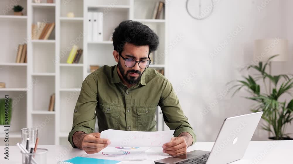 A young Indian male businessman is sitting at a desk trying to work, checking schedules and getting frustrated that something is not working out. Headache, fatigue, burnout.