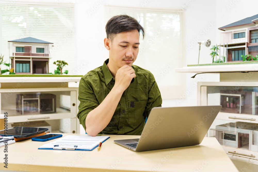 Asian businessman works alone at his desk, surrounded by a laptop, paperwork, tablet, and calculator, focusing on home and land mortgage agreements in a modern house model. Asian businessman works alone at his desk, surrounded by a laptop, paperwork, tablet, and calculator, focusing on home and land mortgage agreements in a modern house model.