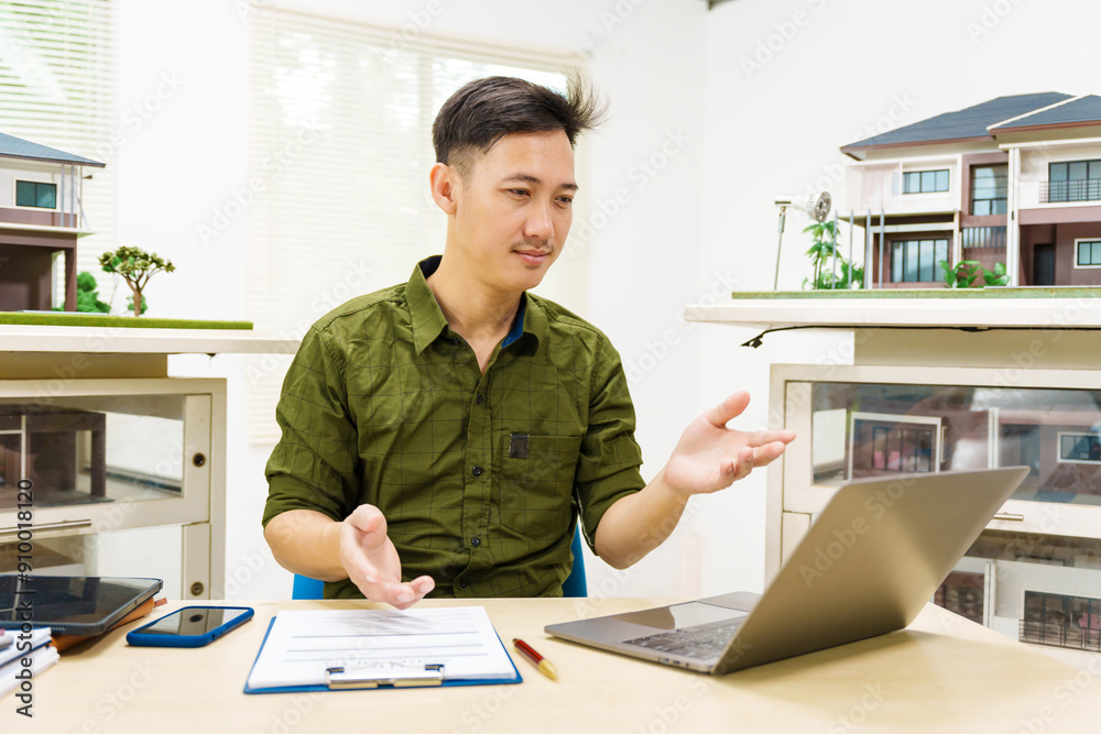 Asian businessman works alone at his desk, surrounded by a laptop, paperwork, tablet, and calculator, focusing on home and land mortgage agreements in a modern house model. Asian businessman works alone at his desk, surrounded by a laptop, paperwork, tablet, and calculator, focusing on home and land mortgage agreements in a modern house model.