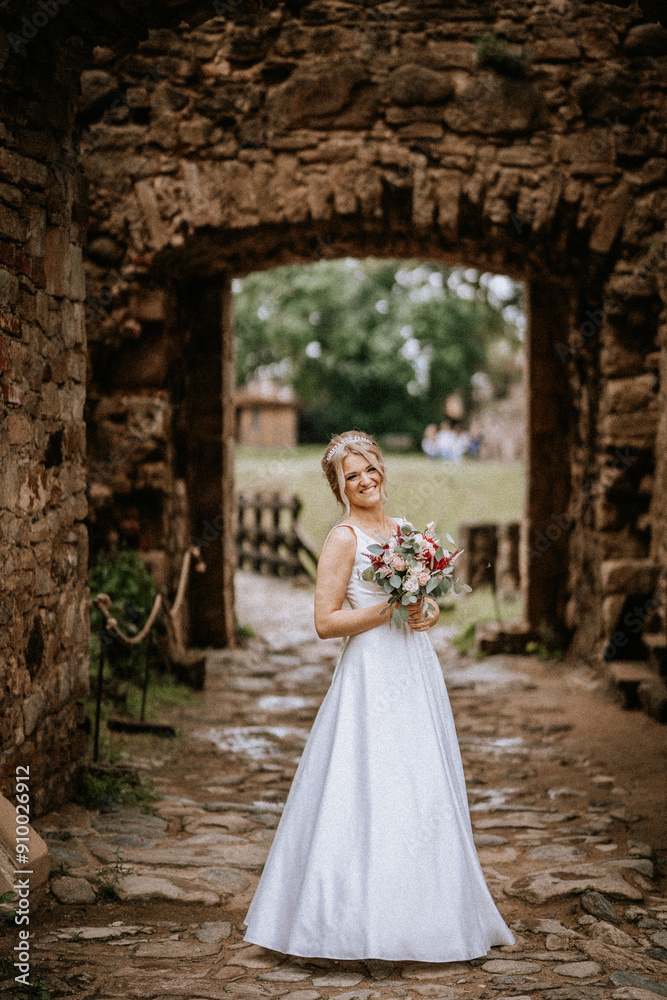 Obraz premium Cesis, Latvia - July 12, 2024 - A smiling bride in a white gown stands holding a bouquet in a stone archway, with a rustic and historic background.