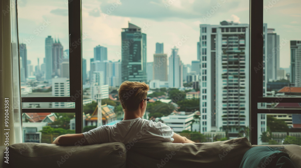A man sits on a sofa, taking in the panoramic city skyline through a large window, suggesting a moment of reflection or relaxation. A man sits on a sofa, taking in the panoramic city skyline through a large window, suggesting a moment of reflection or relaxation.