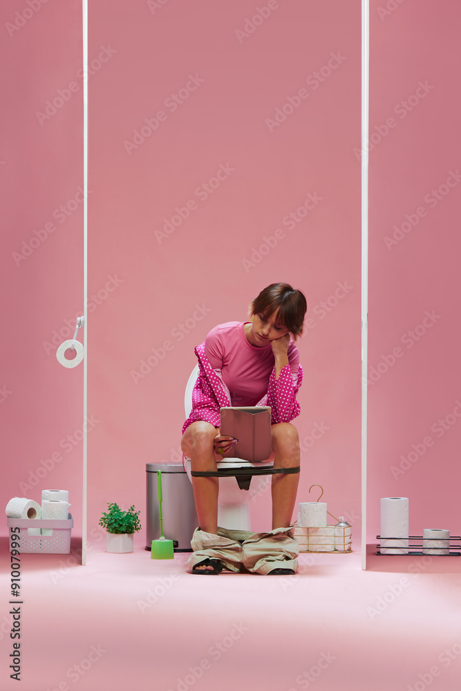 Obraz premium Young girl in pink clothes, with short hair sitting on toilet and looking on tablet with bored face against pink background. Concept of sport, game, match event, human emotions