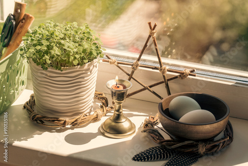 Home spring altar on windowsill with bowl with eggs, the first greens in the pot, candle and birds feathers under the sun's rays, selected focus. photo