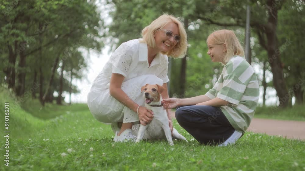 Blonde woman and daughter is walking together along pathway in parkland full of green foliage. Mother and little girl bonding time during weekend with dog, strolling and playing with dog on grass Blonde woman and daughter is walking together along pathway in parkland full of green foliage. Mother and little girl bonding time during weekend with dog, strolling and playing with dog on grass