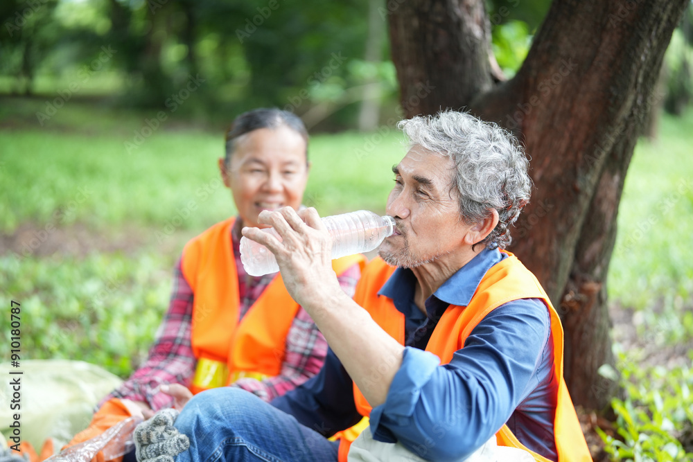 Fototapeta premium older adult husband and wife in safety vest resting under a tree and drinking water,asian senior couple spend holiday time doing volunteer activity collecting and sorting waste in the park