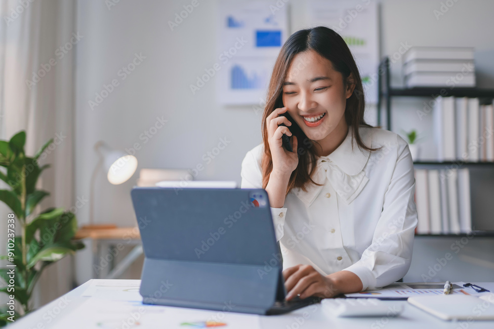 Asian businesswoman is smiling while talking on the phone and using her tablet, looking busy at her desk Asian businesswoman is smiling while talking on the phone and using her tablet, looking busy at her desk