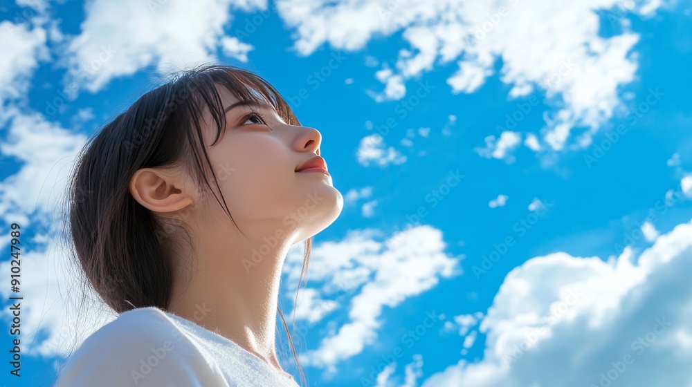 A young Asian woman gazes upward, embracing the beauty of a clear blue sky filled with fluffy white clouds. A young Asian woman gazes upward, embracing the beauty of a clear blue sky filled with fluffy white clouds.