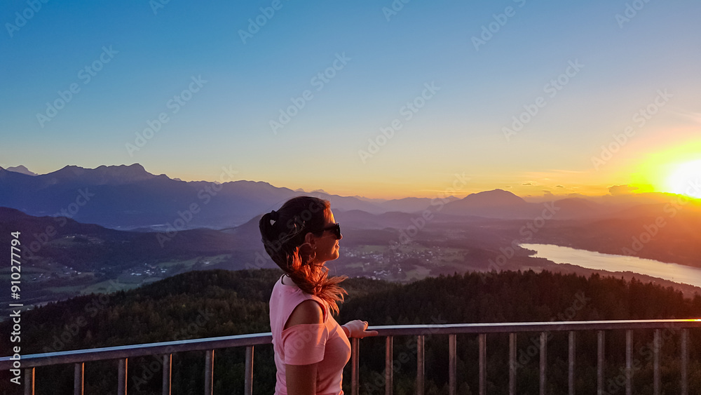 Fototapeta premium Happy woman with panoramic view of lake Woerthersee in summer during sunset seen from observation tower Pyramidenkogel, Carinthia, Austria. Romantic atmosphere surrounded by mountains of Austrian Alps