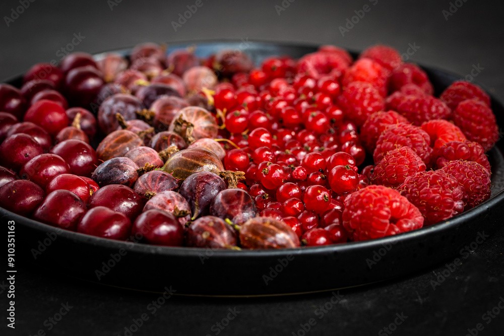 A mixture of red summer berries on black plate, angle view. Tasty fresh ripe red currant, raspberry, gooseberry, cherry berries. Healthy food texture on dark background. A mixture of red summer berries on black plate, angle view. Tasty fresh ripe red currant, raspberry, gooseberry, cherry berries. Healthy food texture on dark background.
