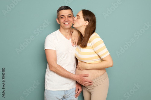 Romantic couple expecting baby husband hugging his lovely pregnant wife touching her bare belly pregnant Caucasian woman kissing her loving man cheek isolated over turquoise background