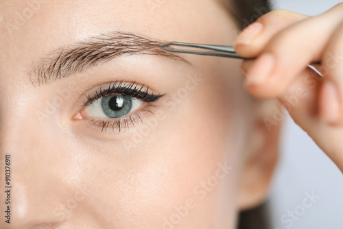 Young woman plucking eyebrow with tweezers on light background, closeup