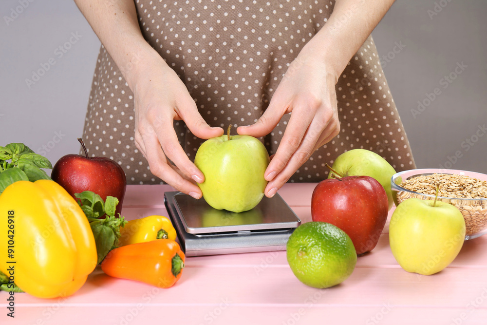 Fototapeta premium Woman weighing apple on kitchen scale at pink wooden table, closeup