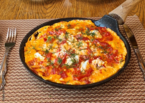 An omelette with tomatoes and fresh herbs is fried in large frying pan over fire