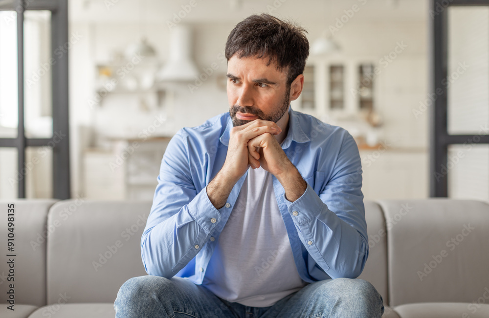 A man with a beard, wearing a blue shirt and jeans, sits on a grey couch in a living room. He has his hands clasped together in front of his chin and is looking off to the side thoughtfully. A man with a beard, wearing a blue shirt and jeans, sits on a grey couch in a living room. He has his hands clasped together in front of his chin and is looking off to the side thoughtfully.