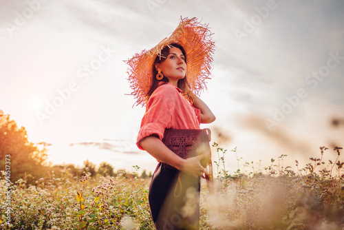 Portrait of young stylish woman walking in blooming buckweat field wearing straw hat holding leather purse photo
