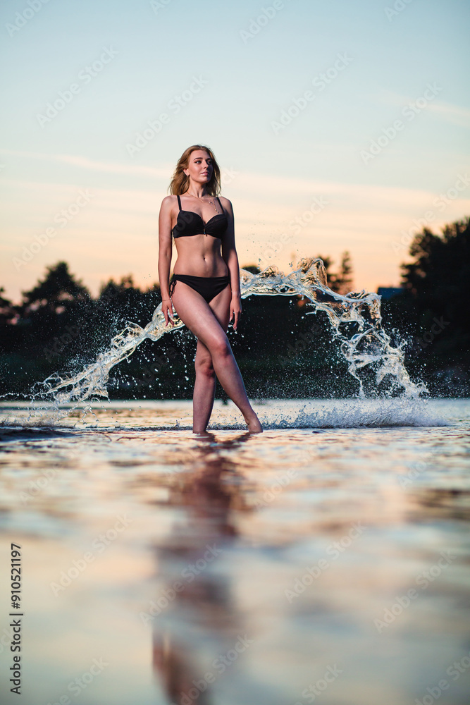 Young slender girl in a black swimsuit posing in the water at sunset at the background of water flying in the air