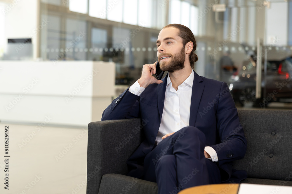 Salesperson in suit having phone conversation while sitting on sofa in contemporary car showroom Salesperson in suit having phone conversation while sitting on sofa in contemporary car showroom