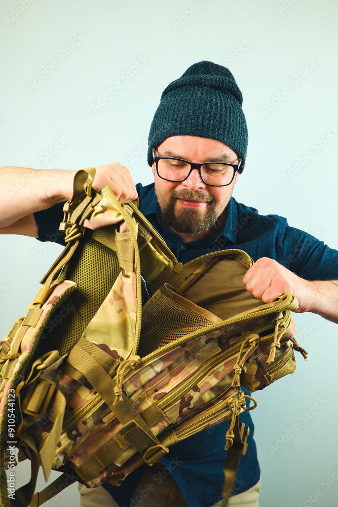 Portrait of handsome bearded man with beanie and glasses looking into camo military backpack, isolated on white background