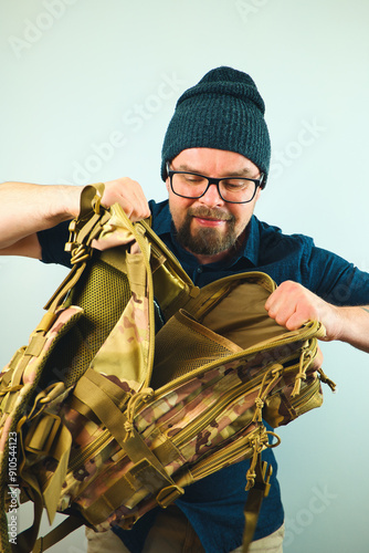 Obraz na płótnie Portrait of handsome bearded man with beanie and glasses looking into camo military backpack, isolated on white background w salonie