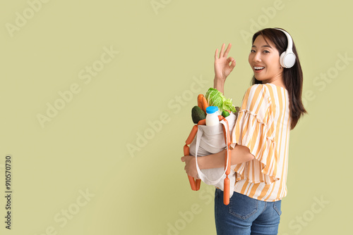 Young Asian woman in headphones with shopper bag full of products showing OK gesture on green background photo