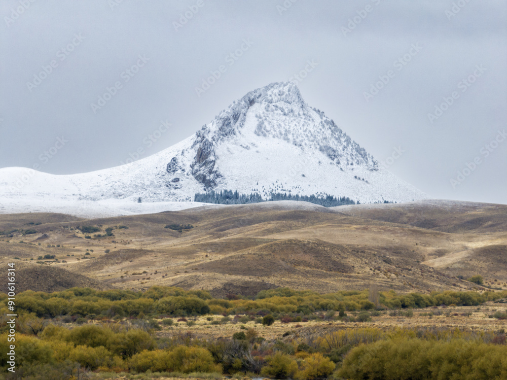 Snow-covered peak rising from an arid landscape with sparse vegetation, under a cloudy sky. Snow-covered peak rising from an arid landscape with sparse vegetation, under a cloudy sky.