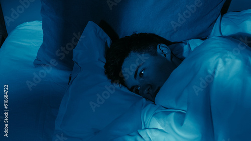 Fotografie Exhausted man experiencing loneliness insomnia lying awake in bed under dim blue lighting