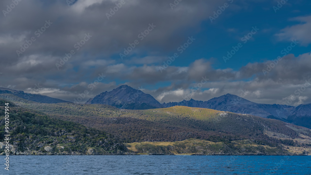 A picturesque mountain range of the Andes against a blue sky and clouds. The forest grows on the slopes of the mountains. In the foreground is the surface of the Beagle Canal. Argentina. A picturesque mountain range of the Andes against a blue sky and clouds. The forest grows on the slopes of the mountains. In the foreground is the surface of the Beagle Canal. Argentina.