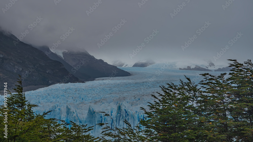 The beautiful Perito Moreno glacier stretches between the mountains to the horizon. An array of blue ice with cracks, sharp peaks. Green trees in the foreground. Cloudy. Fog. Argentina. El Calafate. The beautiful Perito Moreno glacier stretches between the mountains to the horizon. An array of blue ice with cracks, sharp peaks. Green trees in the foreground. Cloudy. Fog. Argentina. El Calafate.