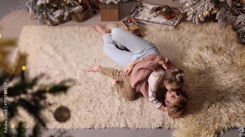 A young mother and daughter 5-6 years old in casual clothes lie on a soft carpet in a cozy, and decorated Christmas living room at home against the backdrop of a Christmas tree.