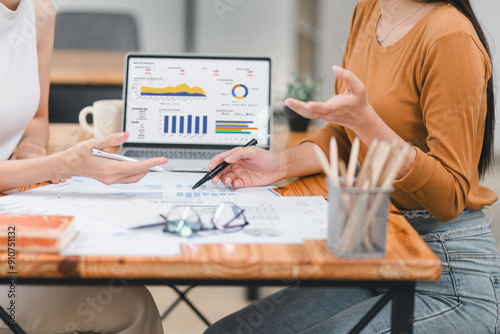 Two professionals analyze business data using laptop displaying sales dashboard and various printed charts on desk. photo