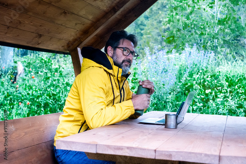 One man with yellow jacker opening metal bottle of coffee sitting ona wooden table at the mountain waiting to work on laptop with roaming data connection. Male mature people in outdoor leisure photo