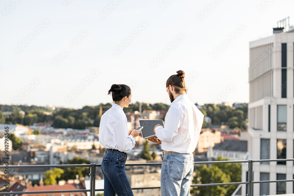 Business colleagues standing on rooftop, discussing project using technology devices. Urban cityscape view in background provides professional setting. Teamwork and collaboration concepts represented. Business colleagues standing on rooftop, discussing project using technology devices. Urban cityscape view in background provides professional setting. Teamwork and collaboration concepts represented.
