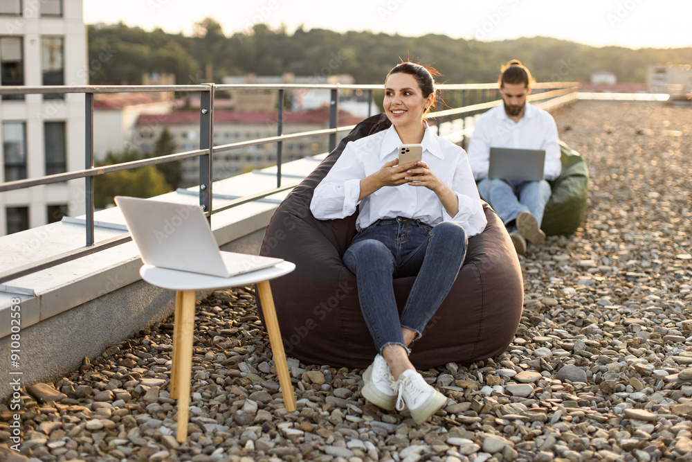 Two people working remotely on rooftop terrace using laptops and smartphones. Outdoor workspace promoting relaxation and productivity, surrounded by urban landscape. Two people working remotely on rooftop terrace using laptops and smartphones. Outdoor workspace promoting relaxation and productivity, surrounded by urban landscape.
