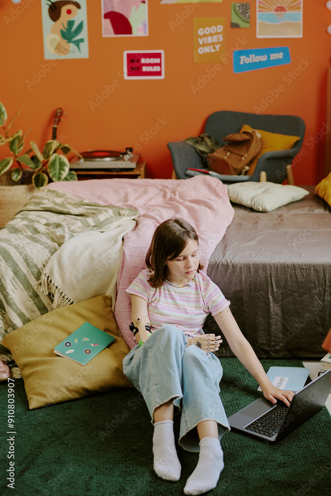 Obraz premium Vertical shot of teenager with disability sitting on floor in her cozy bedroom and studying via laptop, copy space