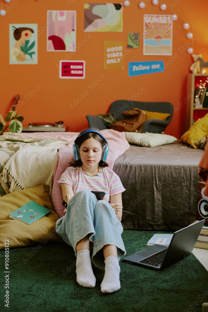 Obraz premium Vertical shot of teen girl with disability in headphones choosing playlist on smartphone while sitting on floor against her bed and doing homework