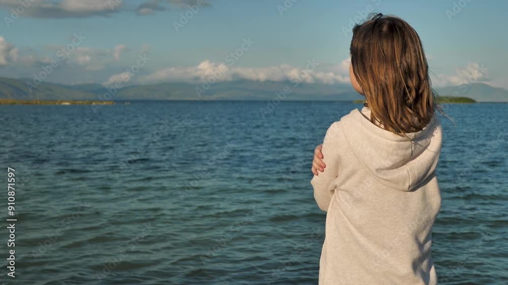 One girl 9 years old standing on the shore of a picturesque racing lake facing the water and admiring the sunset. Teenage girl looking at the sea horizon at sunset. Copy space, view from behind