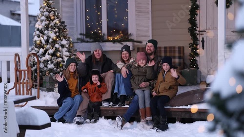 A large family in winter clothes and sweaters poses on the porch of a private country house on Christmas Day against the backdrop of the Christmas tree. The concept of holiday and togetherness.