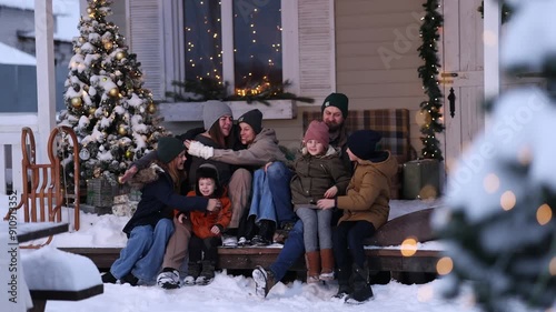 A large family in winter clothes and sweaters poses on the porch of a private country house on Christmas Day against the backdrop of the Christmas tree. The concept of holiday and togetherness.