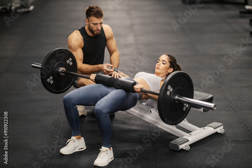 Muscular sportswoman doing hip thrusts on bench with barbell and her coach is motivating her. photo